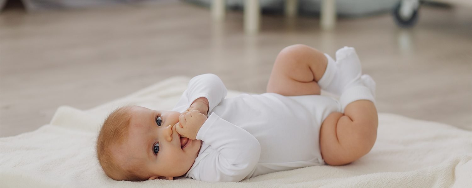 A smiling baby in a white bodysuit lying on a soft cream blanket