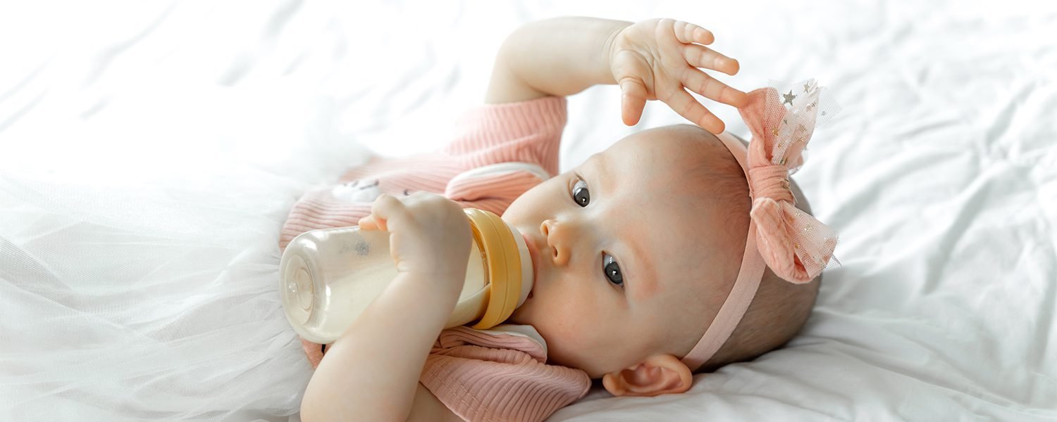 A baby in a pink outfit and headband lying on a white blanket, holding a bottle